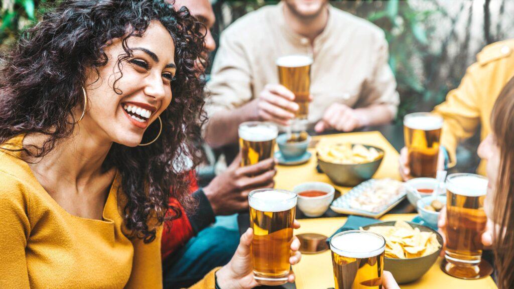 Bild einer fröhlichen Gruppe von Freunden, die zusammen in einem Außenbereich sitzen und Bier trinken. Eine junge Frau mit lockigem Haar und gelbem Oberteil lacht, während sie ihr Bierglas hebt, umgeben von Freunden mit weiteren Biergläsern. Die Szene betont die Freude und Geselligkeit, die Biergeschenke mit sich bringen können