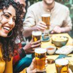 Bild einer fröhlichen Gruppe von Freunden, die zusammen in einem Außenbereich sitzen und Bier trinken. Eine junge Frau mit lockigem Haar und gelbem Oberteil lacht, während sie ihr Bierglas hebt, umgeben von Freunden mit weiteren Biergläsern. Die Szene betont die Freude und Geselligkeit, die Biergeschenke mit sich bringen können