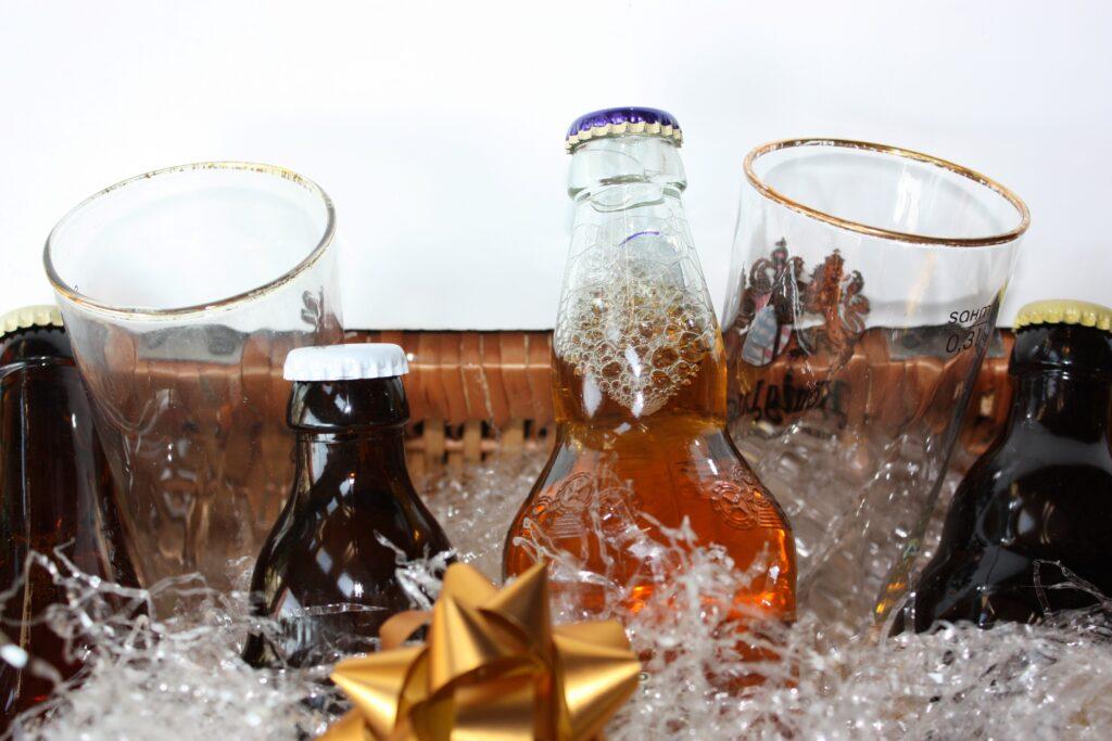 Stillleben von Bierflaschen und -gläsern, kunstvoll arrangiert mit einer goldenen Schleife und künstlichem Schnee, symbolisch für Biergeschenke zur Feierzeit. Die Flaschen und Gläser sind teilweise gefüllt und von funkelndem Konfetti umgeben, was eine festliche und einladende Atmosphäre schafft